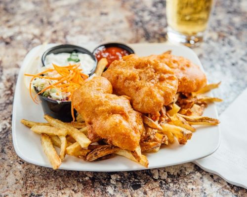 A plate of crispy fish and chips with coleslaw and dipping sauces, served with a glass of beer in the background.