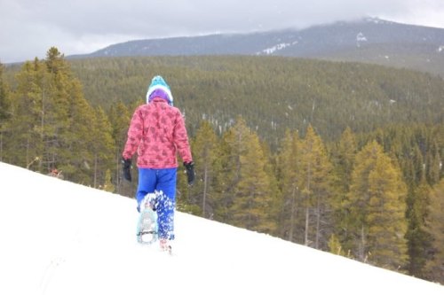 A child in a pink jacket and blue pants walks through snow, surrounded by evergreen trees and mountains in the background.