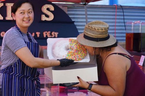 A vendor hands a large, colorful donut to a customer at a food festival, both smiling and enjoying the moment.