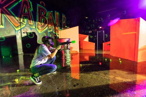 A person crouches with a paintball gun in a colorful arena, aiming at targets amidst vibrant graffiti and neon lights.
