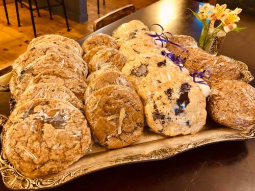 A silver platter filled with assorted wrapped cookies, including oatmeal and chocolate chip, beside a small flower vase.