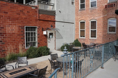 A courtyard with tables and chairs, surrounded by brick buildings and greenery, featuring a small fountain.