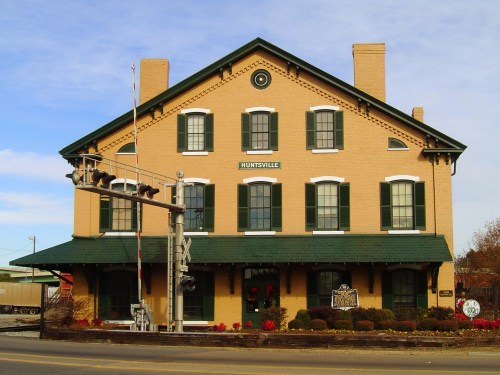 Historic brick building with green trim, labeled "Huntsville," near a railroad crossing and decorated with plants.