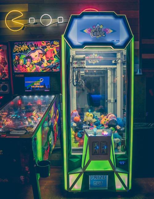 A brightly lit claw machine filled with plush toys, next to a colorful pinball machine in a retro arcade setting.