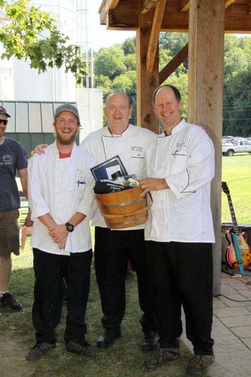 Three chefs in white coats stand together outdoors, smiling and holding a basket, with trees and a guitar in the background.