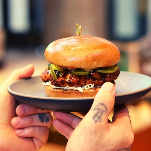 A person holds a plate with a grilled chicken sandwich topped with pickles and a toothpick, on a soft bun.