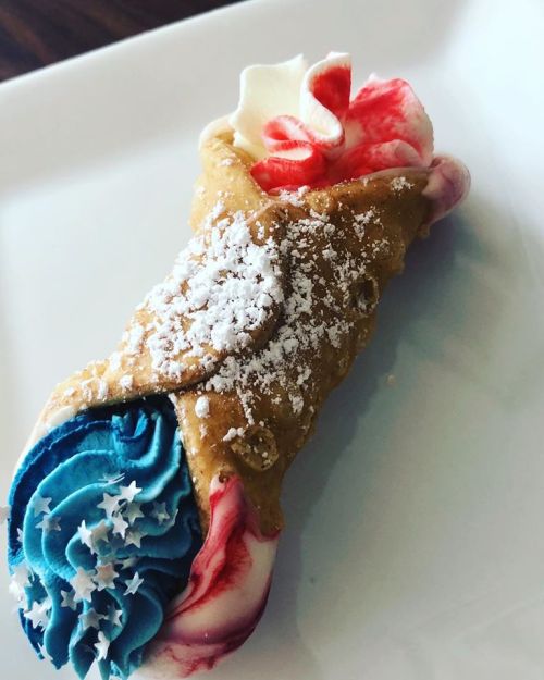 A colorful dessert cannoli with red, white, and blue frosting, topped with powdered sugar and star sprinkles.