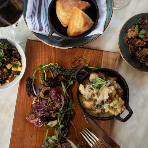 A wooden platter with assorted meats, gnocchi in sauce, and sides, accompanied by cornbread and small bowls of vegetables.