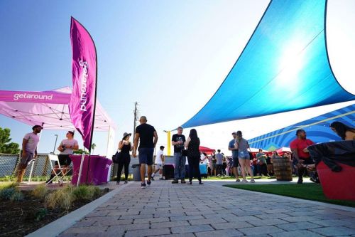 A vibrant outdoor event with colorful tents, people mingling, and shaded areas under blue and pink canopies.