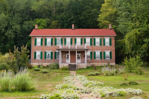 A large brick house with a red roof, green shutters, and a front porch, surrounded by a garden and trees.