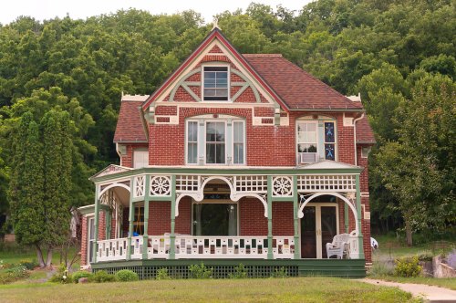 A charming brick house with a large porch, intricate woodwork, and surrounded by lush greenery.