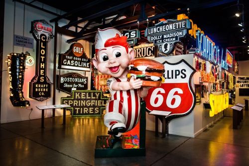 A vintage diner mascot holding a burger stands in front of colorful retro signs in a museum exhibit.