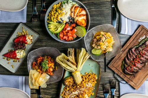A vibrant spread of various dishes, including grilled meats, salads, and corn, arranged on wooden and ceramic plates.