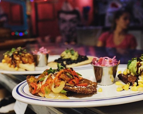 A plate of colorful tacos with toppings, served with a side of pickled onions and corn, in a vibrant restaurant setting.