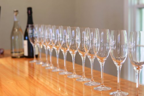 A row of sparkling wine glasses on a wooden bar, with bottles of champagne in the background.