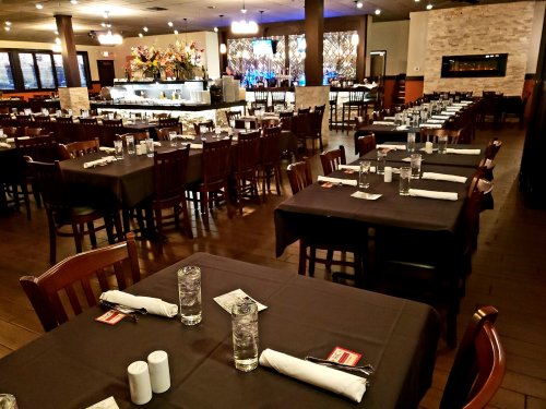 A spacious restaurant with neatly set tables, dark tablecloths, and a bar area in the background, ready for diners.