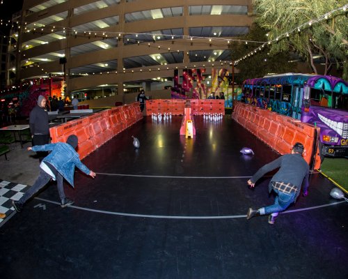 Two people play a bowling game outdoors, with colorful lights and a festive atmosphere in the background.