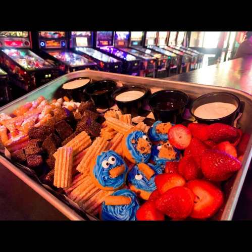 A tray of assorted desserts including churros, cookies, and strawberries, with dipping sauces, set against arcade machines.