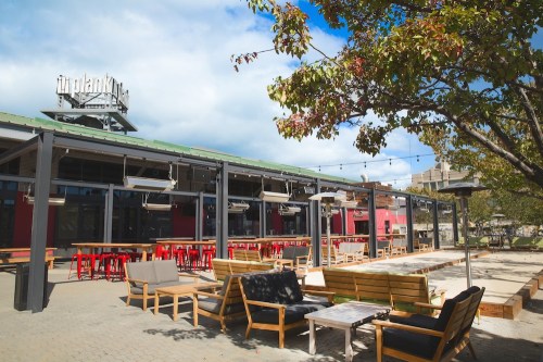 Outdoor seating area of a modern restaurant with wooden furniture, string lights, and a cloudy blue sky.