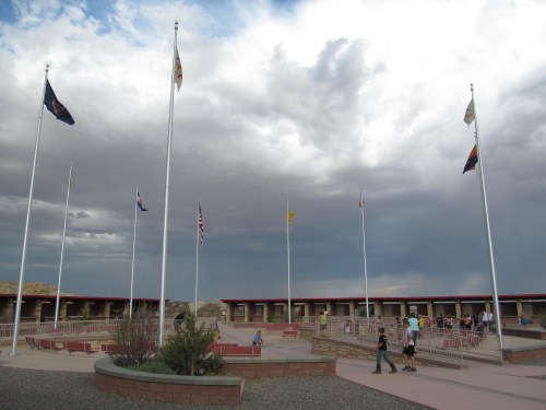 A plaza with multiple flags under a cloudy sky, featuring people walking and sitting around the area.