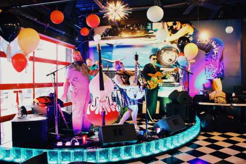 A lively band performs on stage with colorful balloons and a retro car backdrop in a vibrant venue.