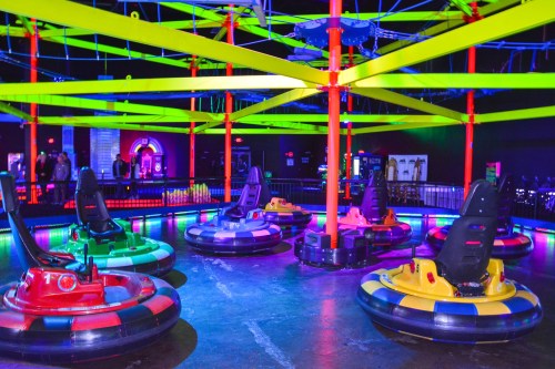 Colorful bumper cars are arranged in a circular arena with vibrant neon lights and a colorful overhead structure.
