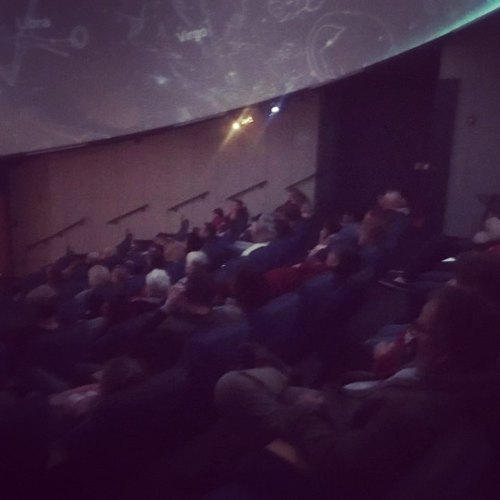 Audience seated in a planetarium, watching a starry projection on the dome ceiling.