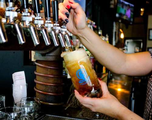 A bartender pours a frothy drink from a tap into a glass at a bar.