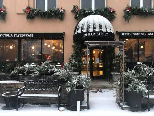 A cozy café entrance adorned with holiday decorations, surrounded by snow-covered plants and a bench.
