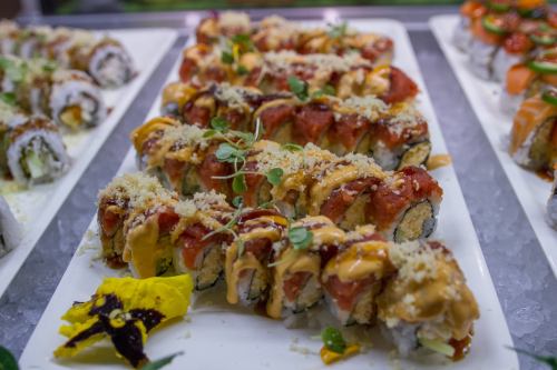 A platter of colorful sushi rolls topped with sauces, crunchy bits, and garnishes, displayed on a white plate.