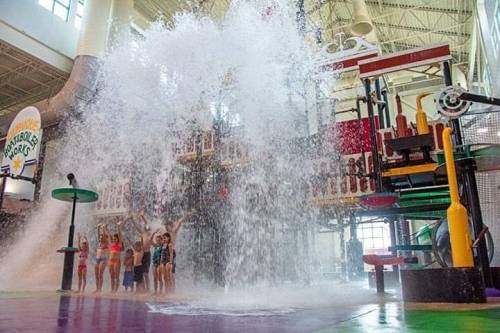 Children play joyfully under a large water splash at an indoor water park, surrounded by colorful structures.
