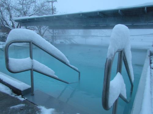 A snowy scene featuring a steaming outdoor pool with snow-covered railings and a misty atmosphere.
