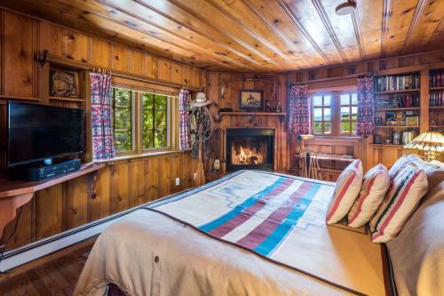 Cozy wooden bedroom with a fireplace, large windows, and a bed adorned with decorative pillows and a colorful blanket.