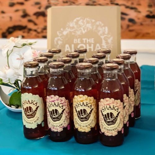 A display of Shaka beverages in glass bottles, featuring flavors like pineapple and ginger, arranged on a blue table.