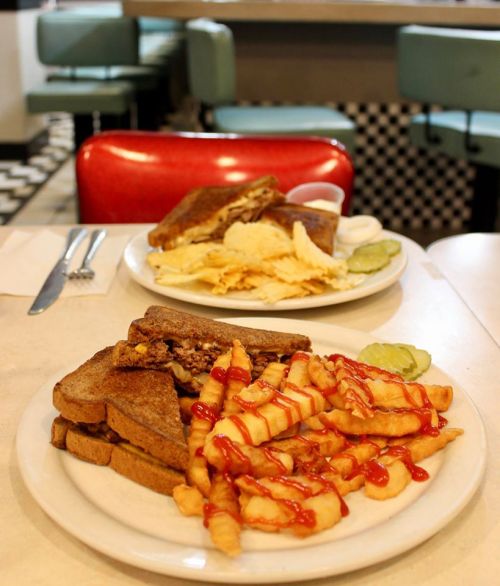 A plate with a grilled sandwich, crinkle-cut fries drizzled with ketchup, and pickles, set in a retro diner.