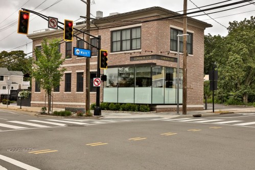 A brick building at an intersection with traffic lights and street signs, surrounded by trees and a clear sky.