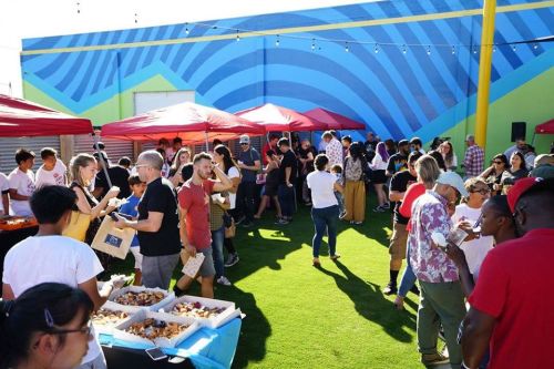 A lively outdoor market scene with people mingling, food stalls, and colorful murals in the background.