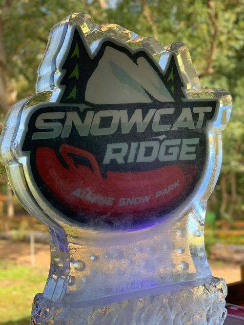 A clear ice sculpture featuring the logo of Snowcat Ridge, an alpine snow park, with mountains and trees in the design.