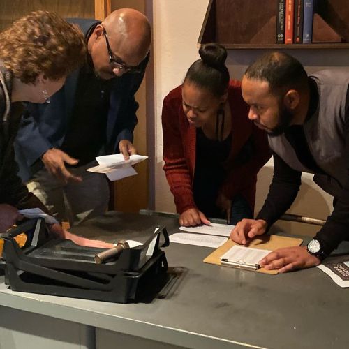 A group of four people gathered around a table, examining documents and discussing something intently.