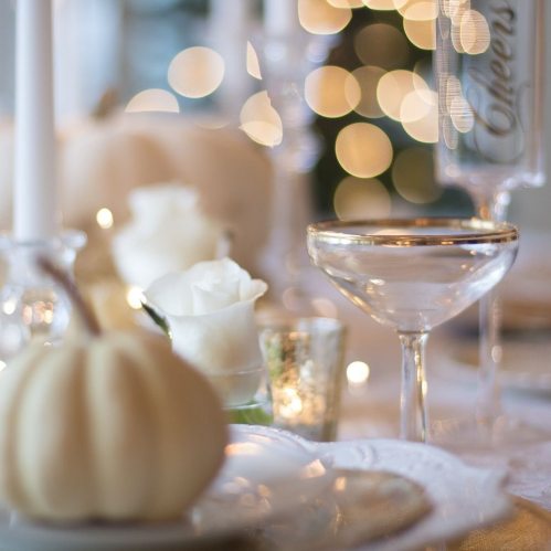 A beautifully set table with white roses, a gold-rimmed glass, and small white pumpkins, illuminated by soft lights.