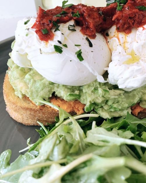A toasted slice of bread topped with smashed avocado, two poached eggs, and sun-dried tomatoes, served with arugula.