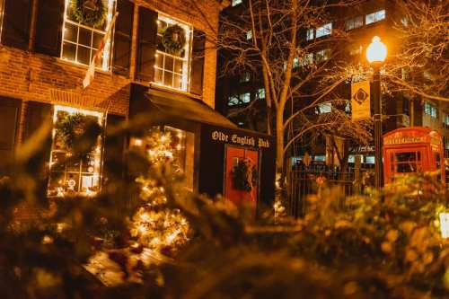 A cozy pub adorned with holiday lights and decorations, surrounded by city buildings and a red telephone booth.