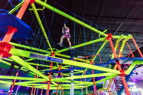 A child navigates a colorful indoor ropes course with bright neon structures and safety harnesses.