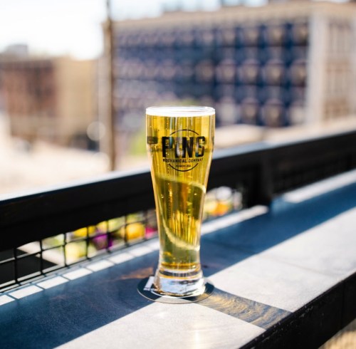 A tall glass of beer sits on a table, with a logo visible, against a blurred city background.