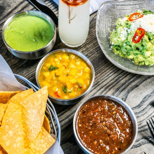 A spread of dips including guacamole, salsa, and mango salsa, served with tortilla chips on a wooden table.