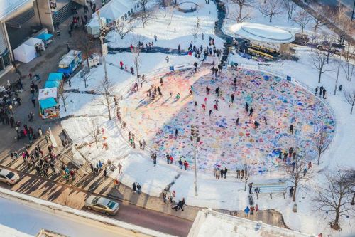 Aerial view of a snowy outdoor area with people skating and colorful artwork on the ice, surrounded by trees and buildings.