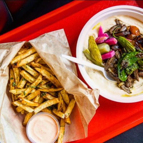 A red tray holds a bowl of grilled vegetables and meat with hummus, alongside a serving of crispy fries and dipping sauce.