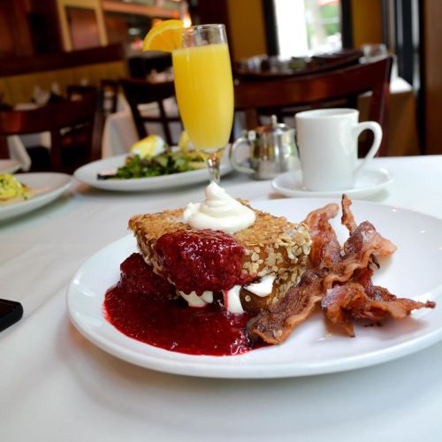 A plate with a dessert topped with whipped cream and raspberry sauce, alongside crispy bacon and a mimosa.