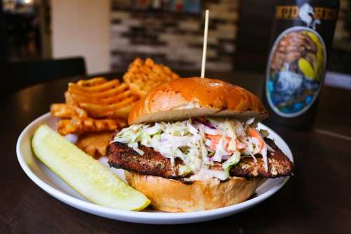 A crispy fish sandwich with coleslaw on a bun, served with waffle fries and a pickle on the side.