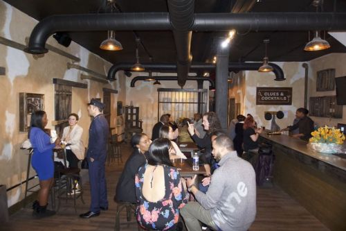 A lively bar scene with people socializing, drinks in hand, and a cozy atmosphere featuring exposed brick and warm lighting.